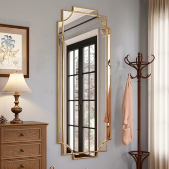Tall gold-framed mirror reflecting a room with a lamp, coat rack, and window.